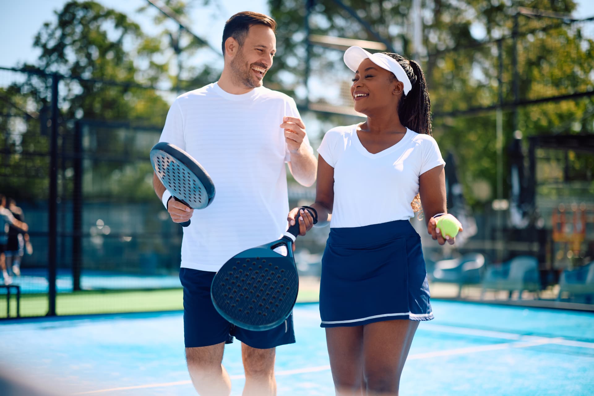 A man and a woman laughing as they walk off a padel court, racquets in hand