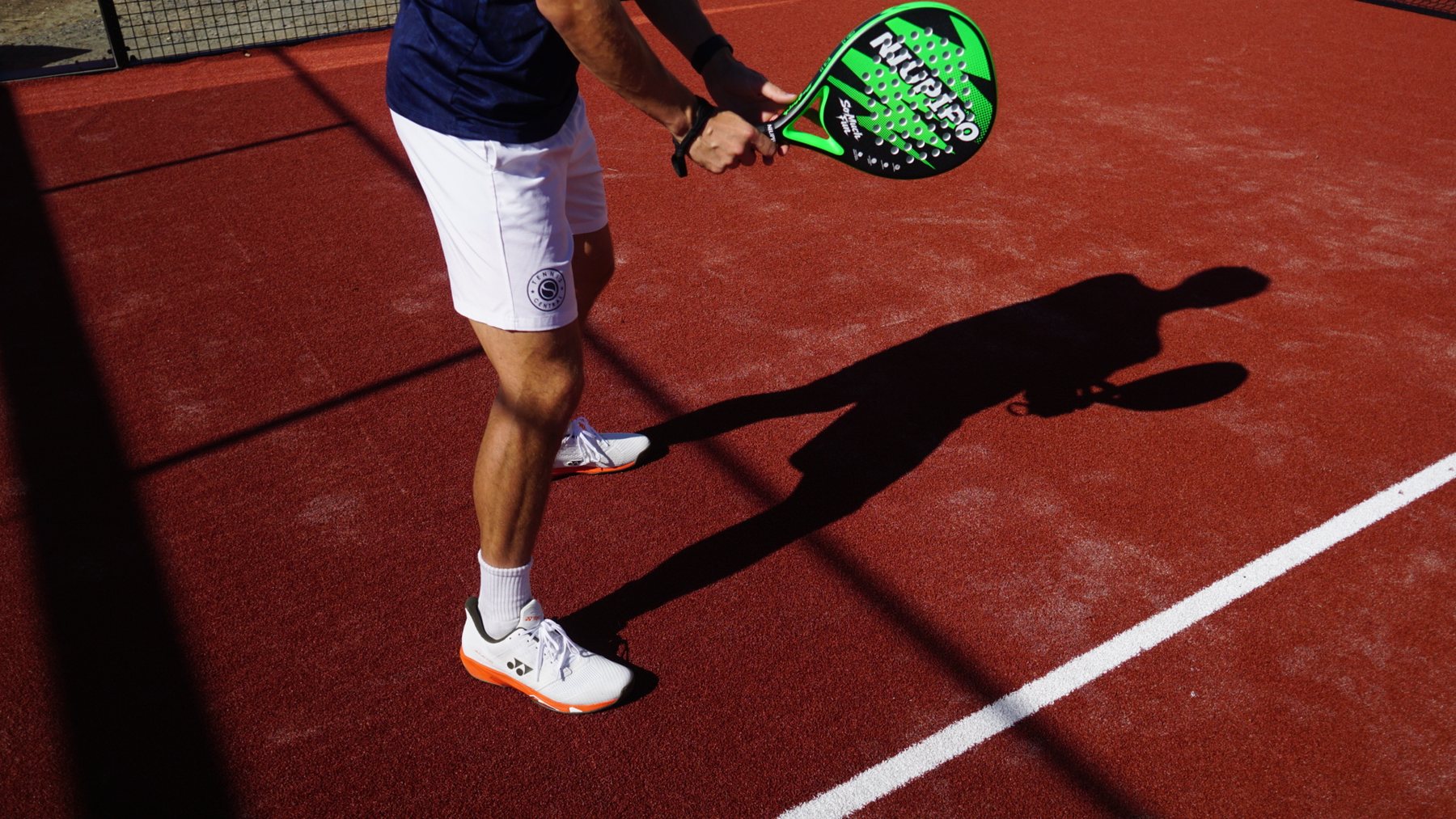 A player in a ready stance on the red court, racquet held in both hands, their shadow stretched across the surface